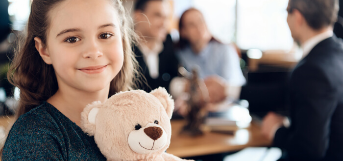 A child holds a teddy bear while an attorney speaks to her parents