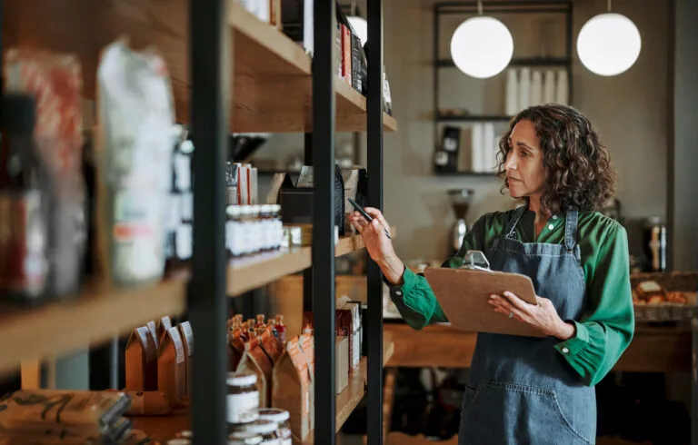Mature female delicatessen owner holding a clipboard while checking inventory of artisanal merchandise on some shelves in her shop